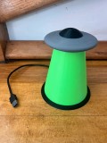 Lampă decorativă cu design inspirat de un OZN și fascicul luminos verde, &icirc;n interiorul căruia se vede silueta unei vaci.